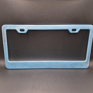 May include: A light blue license plate frame with a textured surface. The frame has a rectangular shape with rounded corners and two mounting holes at the top. The background is black.