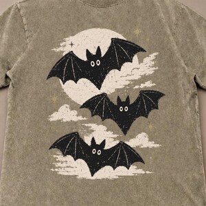 May include: A mineral-washed olive green t-shirt featuring a graphic print of three black bats flying in front of a large white moon and clouds. The design includes small gold stars scattered around the bats and the moon, set against the fabric.