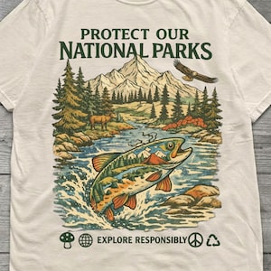 May include: A cream-colored short-sleeve t-shirt featuring a graphic print with the text "PROTECT OUR NATIONAL PARKS" and "EXPLORE RESPONSIBLY". The illustration depicts a jumping fish, mountains, pine trees, an elk, and an eagle.