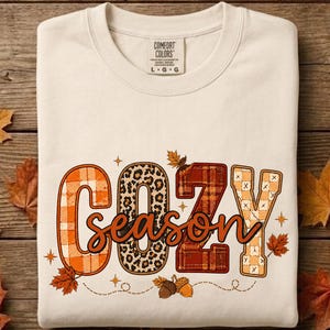 Peut inclure: Un sweat-shirt à col rond couleur crème arborant le texte COZY en grandes lettres majuscules avec des motifs variés incluant du tartan orange, de l'imprimé léopard et du tartan marron. Le mot season est écrit en cursif avec des glands et des feuilles.