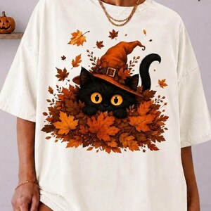 May include: An off-white short-sleeve t-shirt featuring a graphic of a black cat with large yellow eyes wearing an orange witch hat. The cat is nestled in a pile of vibrant orange and brown autumn leaves. The design is centered on the chest.