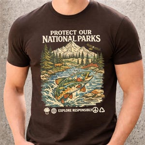 May include: A dark brown short-sleeve t-shirt featuring a graphic print with the text PROTECT OUR NATIONAL PARKS and EXPLORE RESPONSIBLY. The illustration depicts a mountain landscape, a river, a fish, an eagle, and small icons of a mushroom, globe, peace sign, and recycling symbol.