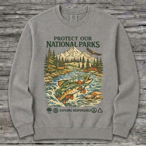 May include: A grey crewneck sweatshirt featuring a graphic print with the text PROTECT OUR NATIONAL PARKS and EXPLORE RESPONSIBLY. The design depicts a mountain landscape, a forest, a flying eagle, and a large fish jumping out of a river.