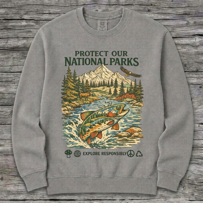 May include: A grey crewneck sweatshirt featuring a graphic print with the text PROTECT OUR NATIONAL PARKS and EXPLORE RESPONSIBLY. The design depicts a mountain landscape, a forest, a flying eagle, and a large fish jumping out of a river.