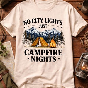 May include: Cream-colored t-shirt featuring a camping scene with a tent, campfire, and mountains. The text reads "NO CITY LIGHTS JUST CAMPFIRE NIGHTS" in a black, bold font. The design evokes a sense of outdoor adventure and nature.