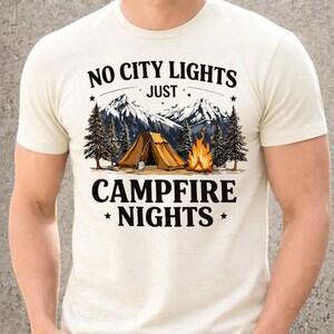 May include: Cream-colored t-shirt with a camping-themed graphic. The design features a tent, campfire, mountains, and pine trees. Text reads "NO CITY LIGHTS JUST CAMPFIRE NIGHTS". The shirt is made of a soft material.