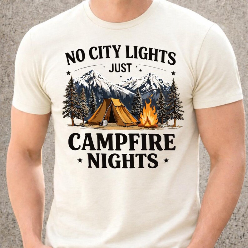 May include: Cream-colored t-shirt with a camping-themed graphic. The design features a tent, campfire, mountains, and pine trees. Text reads "NO CITY LIGHTS JUST CAMPFIRE NIGHTS". The shirt is made of a soft material.
