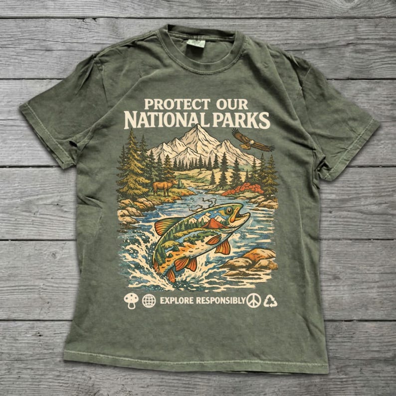 May include: An olive green short-sleeve t-shirt featuring a graphic print of a mountain landscape, a forest, a flying eagle, and a jumping fish. The text reads PROTECT OUR NATIONAL PARKS and EXPLORE RESPONSIBLY, accompanied by small icons.