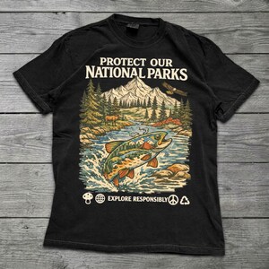May include: A black short-sleeve t-shirt featuring a graphic print with the text PROTECT OUR NATIONAL PARKS. The design depicts a mountain landscape, a forest, an eagle, and a large fish jumping out of water, with EXPLORE RESPONSIBLY below.