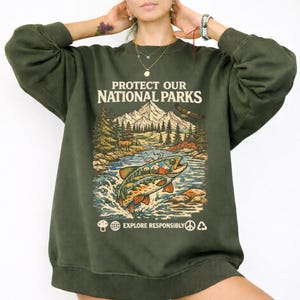 May include: An olive green crewneck sweatshirt featuring a graphic print with the text "PROTECT OUR NATIONAL PARKS" and "EXPLORE RESPONSIBLY". The design depicts a mountain landscape, a forest, an eagle, and a jumping fish in the water.