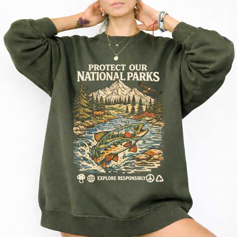 May include: An olive green crewneck sweatshirt featuring a graphic print with the text "PROTECT OUR NATIONAL PARKS" and "EXPLORE RESPONSIBLY". The design depicts a mountain landscape, a forest, an eagle, and a jumping fish in the water.