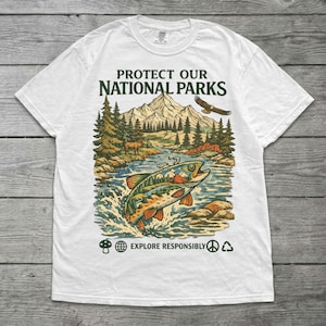 May include: A white t-shirt featuring a graphic print with the text PROTECT OUR NATIONAL PARKS. The design shows a mountain landscape, a forest, a river, a jumping fish, and an eagle. Below, the text EXPLORE RESPONSIBLY with small icons.