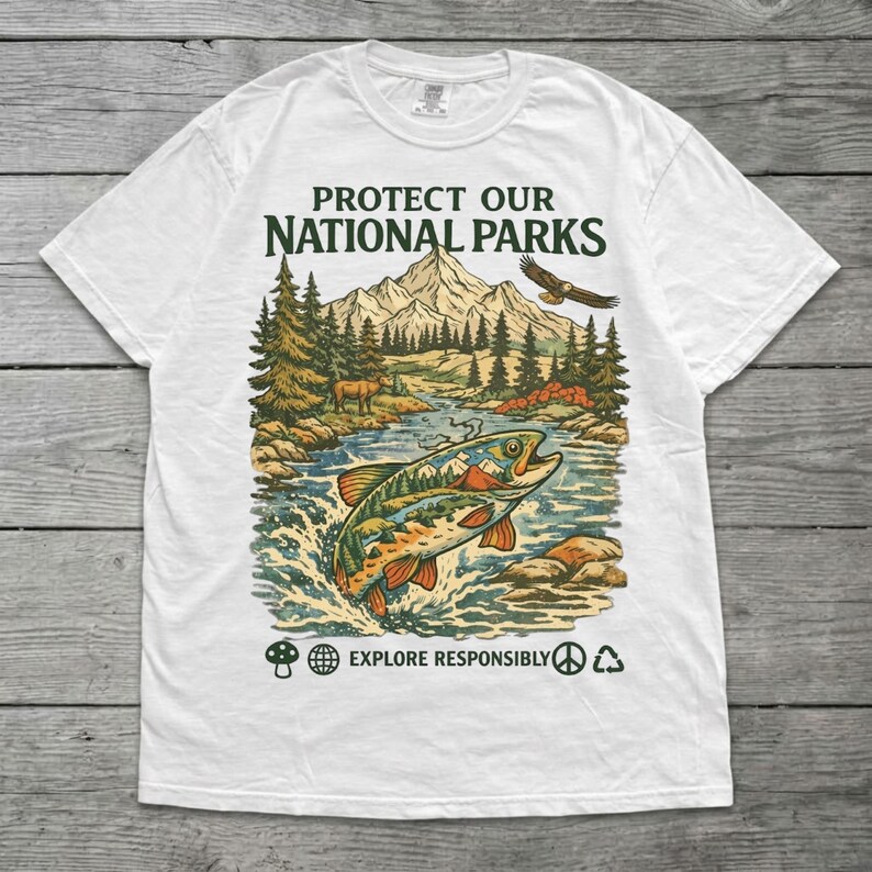 May include: A white t-shirt featuring a graphic print with the text PROTECT OUR NATIONAL PARKS. The design shows a mountain landscape, a forest, a river, a jumping fish, and an eagle. Below, the text EXPLORE RESPONSIBLY with small icons.