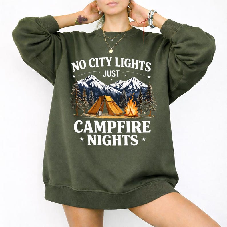 May include: An olive green sweatshirt with the text "NO CITY LIGHTS JUST CAMPFIRE NIGHTS" in white. The graphic features a tent, campfire, mountains, and trees. The sweatshirt is a relaxed fit.