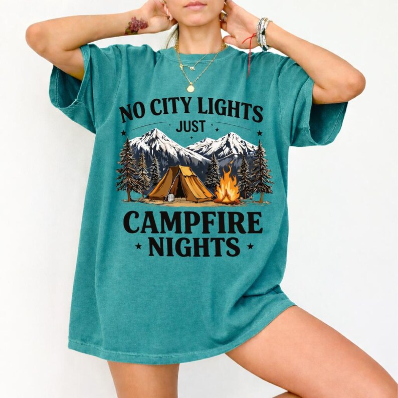 May include: Teal-colored t-shirt with the text "NO CITY LIGHTS JUST CAMPFIRE NIGHTS" above a graphic of a tent, campfire, and mountains. The shirt is a short-sleeved, casual style.