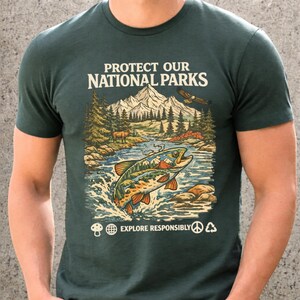 May include: A dark green short-sleeve t-shirt featuring a graphic of a mountain landscape, a river, and a jumping fish. The text reads PROTECT OUR NATIONAL PARKS and EXPLORE RESPONSIBLY, accompanied by icons of a mushroom, globe, peace sign, and recycle symbol.