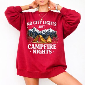 May include: A red sweatshirt with the text "NO CITY LIGHTS JUST CAMPFIRE NIGHTS" in white. The graphic features a camping scene with mountains, trees, a tent, and a campfire. The sweatshirt is long-sleeved.