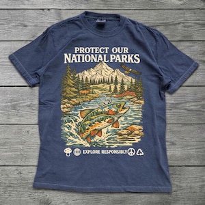 May include: A navy blue short-sleeve t-shirt featuring a graphic print of a mountain landscape, a river, and a fish. The text reads PROTECT OUR NATIONAL PARKS and EXPLORE RESPONSIBLY, accompanied by icons of a mushroom, globe, peace sign, and recycle symbol.