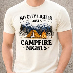 May include: Cream-colored t-shirt with a camping-themed graphic. The design features a mountain range, pine trees, a tent, and a campfire. Text reads "NO CITY LIGHTS JUST CAMPFIRE NIGHTS."