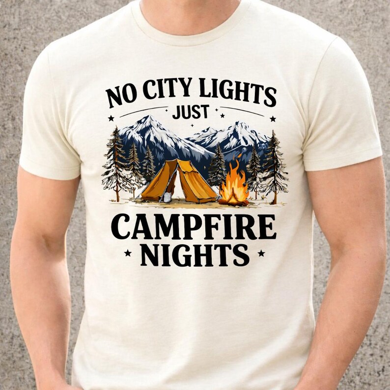 May include: Cream-colored t-shirt with a camping-themed graphic. The design features a mountain range, pine trees, a tent, and a campfire. Text reads "NO CITY LIGHTS JUST CAMPFIRE NIGHTS."