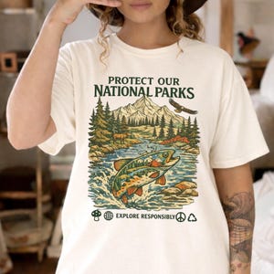 May include: A cream-colored t-shirt featuring a graphic of a mountain landscape, a forest, a deer, and a fish jumping in a river. The text reads PROTECT OUR NATIONAL PARKS and EXPLORE RESPONSIBLY, accompanied by small icons of a mushroom, globe, peace sign, and recycling symbol.