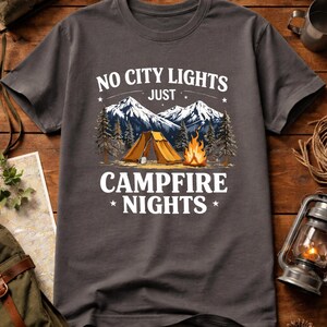 May include: Dark gray t-shirt with a camping graphic. The design features a tent, campfire, and mountains, with the text "NO CITY LIGHTS JUST CAMPFIRE NIGHTS". The shirt is made of a soft material and is suitable for outdoor enthusiasts.