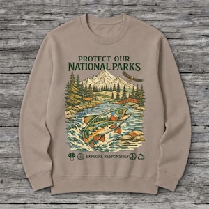 May include: A tan crewneck sweatshirt featuring a graphic print with the text PROTECT OUR NATIONAL PARKS and EXPLORE RESPONSIBLY. The illustration depicts a jumping fish, a mountain range, pine trees, a flying eagle, and a deer.