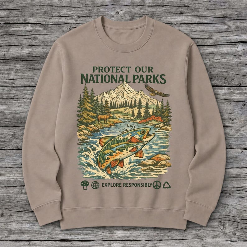 May include: A tan crewneck sweatshirt featuring a graphic print with the text PROTECT OUR NATIONAL PARKS and EXPLORE RESPONSIBLY. The illustration depicts a jumping fish, a mountain range, pine trees, a flying eagle, and a deer.