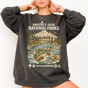 May include: A charcoal gray crewneck sweatshirt featuring a graphic print with the text PROTECT OUR NATIONAL PARKS and EXPLORE RESPONSIBLY. The illustration depicts a mountain landscape, a forest, a flying eagle, and a large jumping fish.
