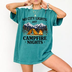 May include: Teal-colored t-shirt with the text "NO CITY LIGHTS JUST CAMPFIRE NIGHTS" above a graphic of a tent, campfire, mountains, and trees. The shirt has short sleeves and a relaxed fit.