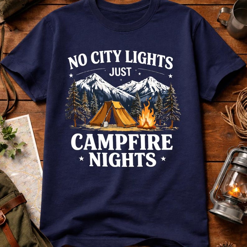 May include: Navy blue t-shirt with a camping scene. The design features a tent, campfire, mountains, and the text "NO CITY LIGHTS JUST CAMPFIRE NIGHTS". The graphic is in white and orange tones.