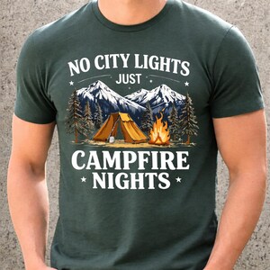May include: Dark green t-shirt with the text "NO CITY LIGHTS JUST CAMPFIRE NIGHTS". The graphic features a mountain range, a tent, a campfire, and pine trees. The design is in white and tan.