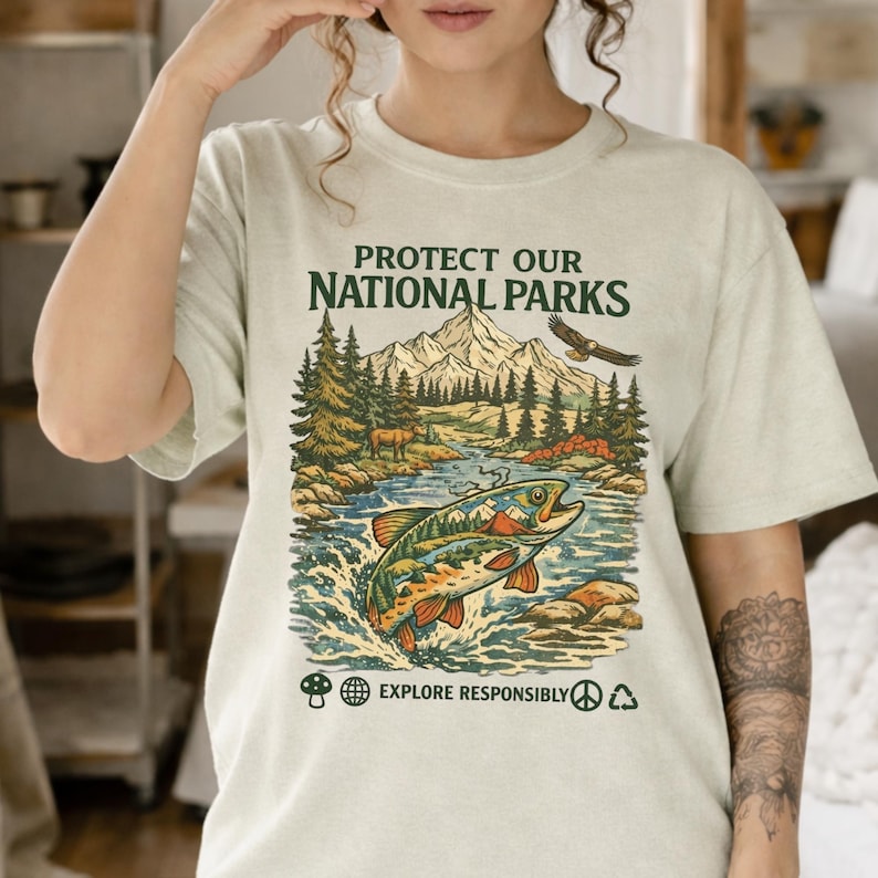 May include: A light beige t-shirt featuring a graphic print with the text "PROTECT OUR NATIONAL PARKS" and "EXPLORE RESPONSIBLY". The illustration depicts a jumping fish, a forest, mountains, an eagle, and a deer, with small icons below.
