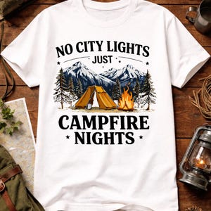 May include: White t-shirt with a camping-themed graphic. The design features a tent, campfire, mountains, and the text "NO CITY LIGHTS JUST CAMPFIRE NIGHTS". The graphic is in shades of brown, orange, and blue.