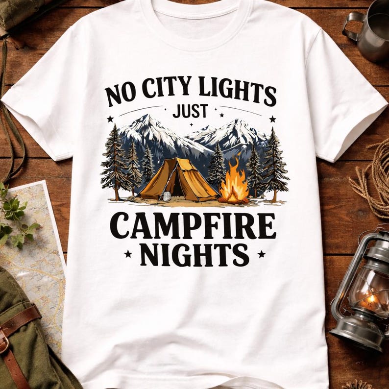 May include: White t-shirt with a camping graphic. The design features a tent, campfire, mountains, and pine trees. Text reads "NO CITY LIGHTS JUST CAMPFIRE NIGHTS".