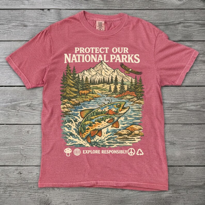 May include: A heathered red short-sleeve t-shirt featuring a graphic print with the text PROTECT OUR NATIONAL PARKS and EXPLORE RESPONSIBLY. The illustration depicts a mountain range, a forest, a flying eagle, and a large fish in a river.