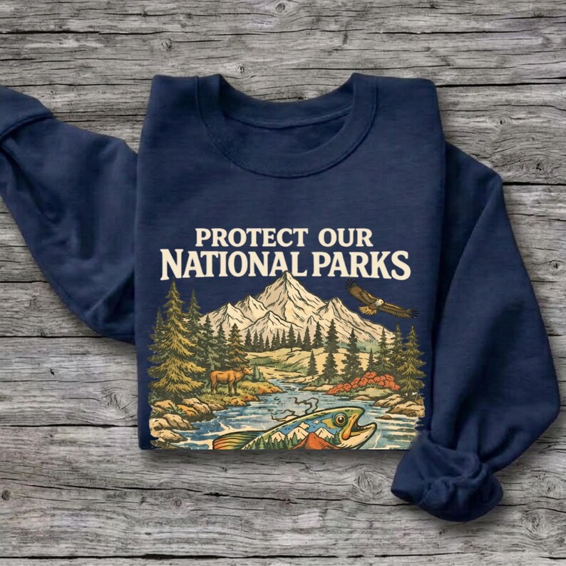 May include: A navy blue long-sleeve sweatshirt featuring a graphic print with the text PROTECT OUR NATIONAL PARKS. The illustration depicts a mountain range, a forest, a river, a deer, a flying eagle, and a large fish in the foreground.