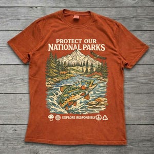 May include: A burnt orange t-shirt featuring a graphic print of a mountain landscape, a river, a fish, and an eagle. The text reads PROTECT OUR NATIONAL PARKS and EXPLORE RESPONSIBLY, accompanied by small icons of a mushroom, globe, peace sign, and recycling symbol.