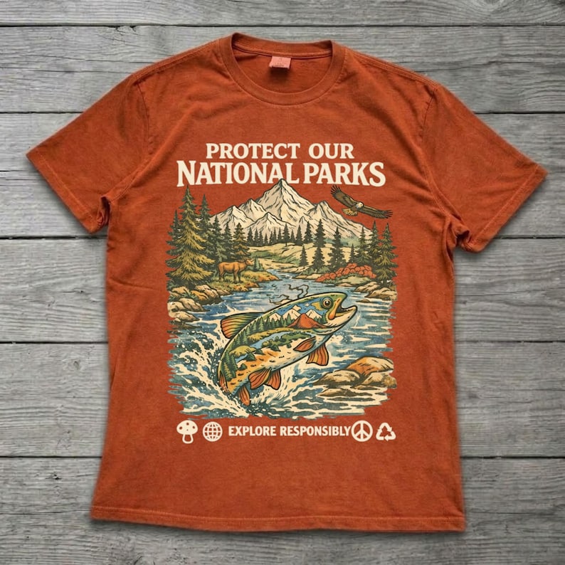 May include: A burnt orange t-shirt featuring a graphic print of a mountain landscape, a river, a fish, and an eagle. The text reads PROTECT OUR NATIONAL PARKS and EXPLORE RESPONSIBLY, accompanied by small icons of a mushroom, globe, peace sign, and recycling symbol.