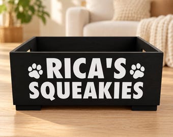 May include: A black rectangular wooden storage crate with cutout handles on the sides. The front features white text that reads RICA'S SQUEAKIES, flanked by a white paw print icon on both the left and right sides of the name.