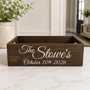 May include: A rectangular dark brown wooden storage crate with cutout handles on the sides. The front panel features white cursive text that reads The Stowe's and the date October 10th 2026 printed in a clean white serif font below the name.