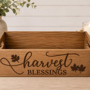 Può includere: Una cassetta rettangolare in legno con finitura marrone medio e maniglie intagliate sui lati. Il pannello frontale presenta la scritta marrone scuro "harvest BLESSINGS" in un carattere decorativo corsivo e stampatello, con foglie d'acero.