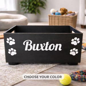May include: A black wooden pet toy box with the name "Buxton" in white script, flanked by white paw prints. The box has a rectangular shape with cut-out handles and sits on small feet. The text "CHOOSE YOUR COLOR" is at the bottom.