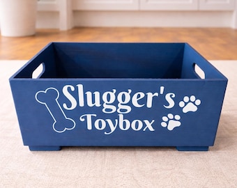 May include: A rectangular navy blue wooden storage bin with cutout handles on the sides. The front features white text that reads Slugger's Toybox, accompanied by a white graphic of a dog bone on the left and two white paw prints on the right.