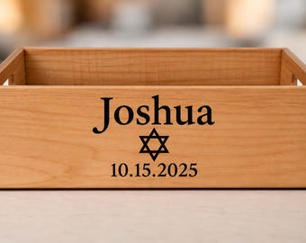 May include: A rectangular light brown wooden storage crate with cutout handles. The front panel features the black printed text Joshua, a Star of David symbol, and the date 10.15.2025 centered on the smooth grain surface.