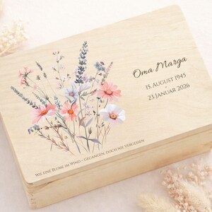 Könnte beinhalten: Eine hellfarbene Holzkiste mit einem Blumendesign mit rosa, weißen und blauen Blumen. Auf der Kiste steht der Name "Oma Marga" und die Daten: 15. August 1945 und 23. Januar 2026. Der Text "WIE EINE BLUME IM WIND - GEGANGEN, DOCH NIE VERGESSEN" ist ebenfalls vorhanden.