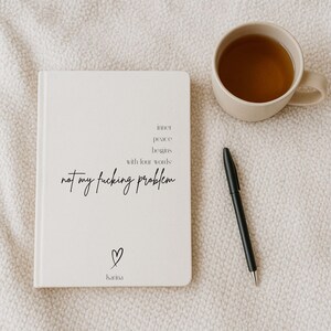 Puede incluir: Un diario de color crema con el texto "inner peace begins with four words not my fucking problem" y un s&iacute;mbolo de coraz&oacute;n. Un bol&iacute;grafo negro y una taza beige llena de t&eacute; est&aacute;n junto al diario sobre una superficie texturizada.