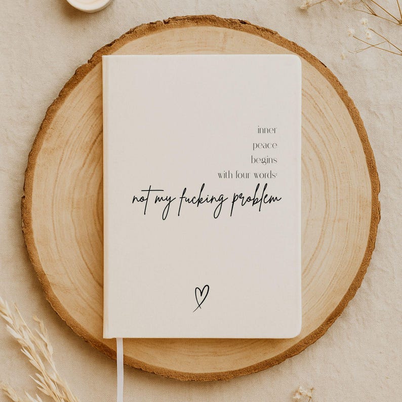 Puede incluir: Un cuaderno color crema con el texto "inner peace begins with four words: not my fucking problem" en escritura negra. Un peque&ntilde;o coraz&oacute;n est&aacute; dibujado en la parte inferior. El cuaderno descansa sobre una rodaja de madera.