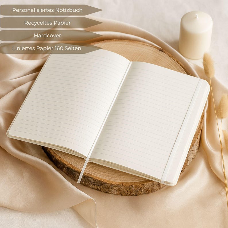 Puede incluir: Un cuaderno rayado abierto con una cubierta blanca y un marcador de cinta descansa sobre una rodaja de madera. El cuaderno est&aacute; rodeado de una tela beige, una vela blanca y plantas secas. El texto en la imagen dice: Cuaderno personalizado, Papel reciclado, Tapa dura, Papel rayado 160 p&aacute;ginas.