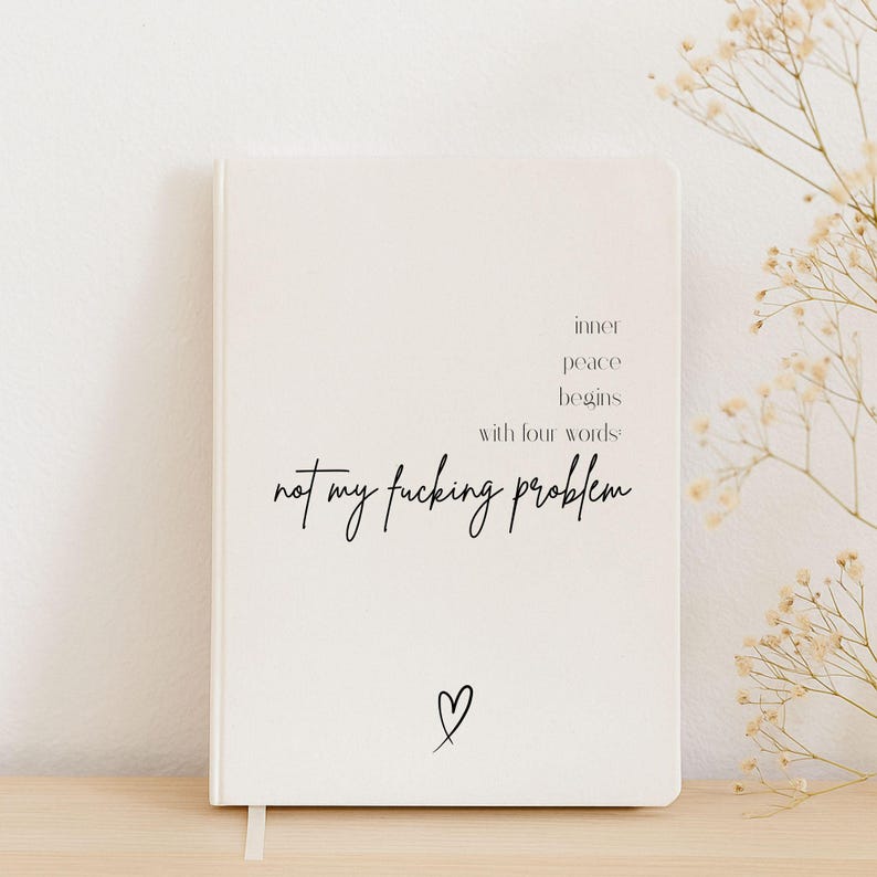 Puede incluir: Un cuaderno color crema con el texto "inner peace begins with four words not my fucking problem" en escritura negra. Un peque&ntilde;o coraz&oacute;n est&aacute; dibujado en la parte inferior. Una cinta blanca como marcap&aacute;ginas es visible.